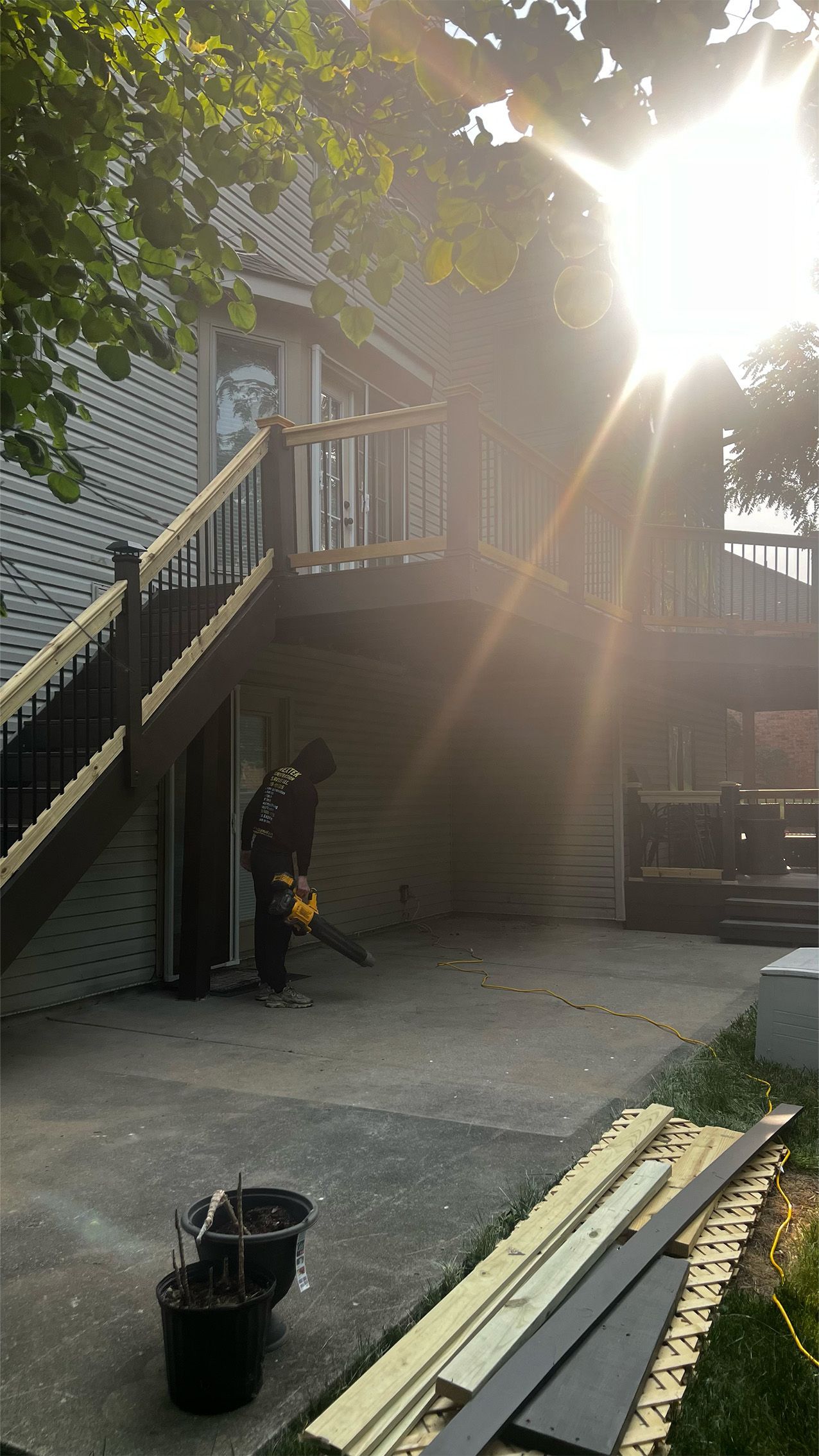 Person using a leaf blower on a concrete patio next to a house with a wooden deck; sunny.