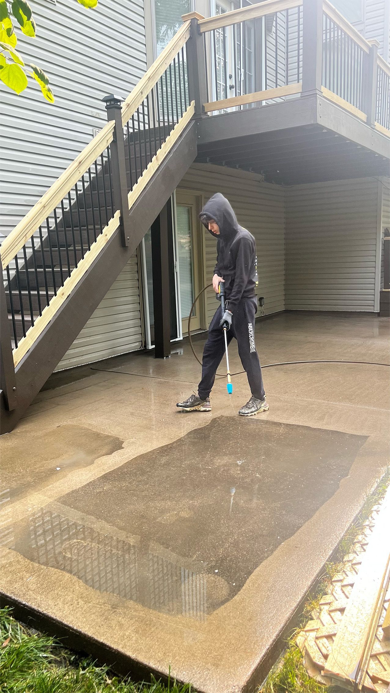 Person in hoodie washes a concrete patio under a wooden deck.