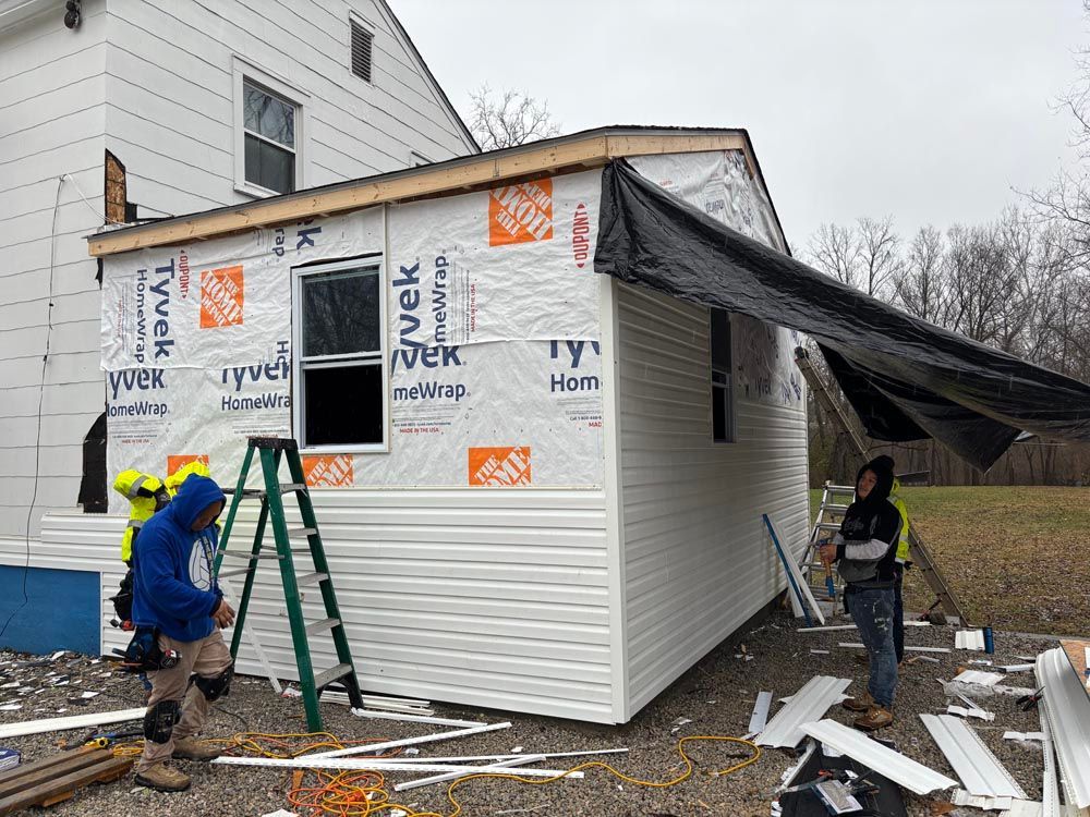 Construction workers installing siding on a white house addition. The addition has new windows, tarp, and Tyvek wrap.