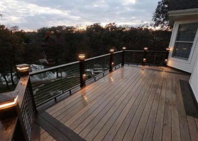 Wooden deck with black railing and lights overlooking a wooded area.