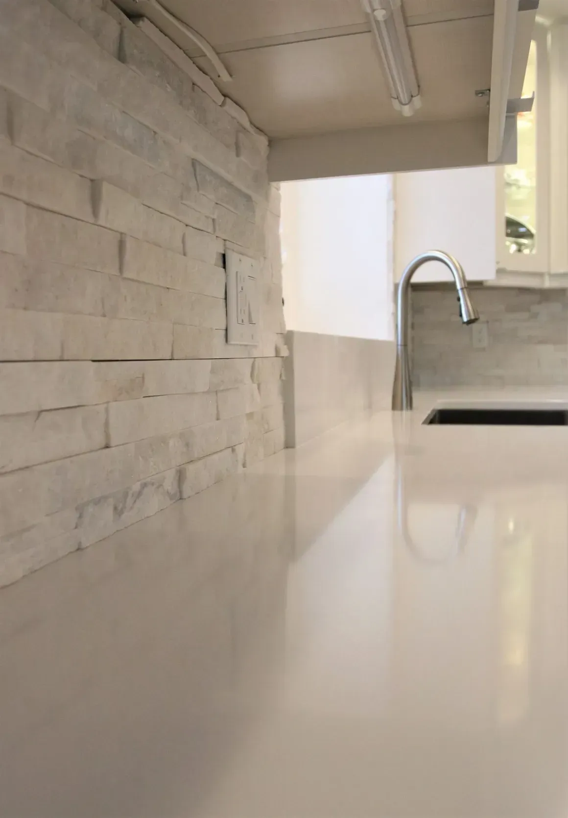 Close-up of a kitchen countertop with a stacked stone backsplash, a modern faucet, and overhead lighting.