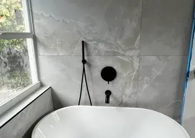 White bathtub in bathroom with gray marble-look wall tiles, black faucet, and window.
