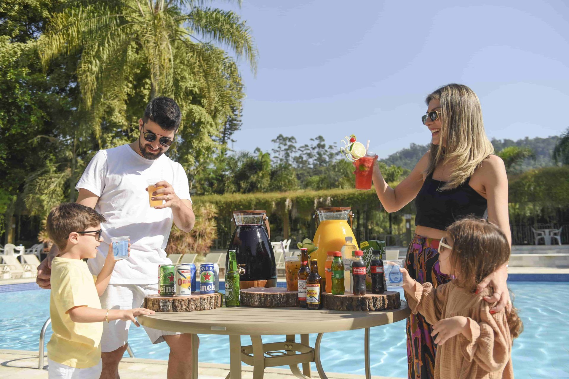 Pessoas à beira da piscina desfrutando de bebidas e petiscos em uma mesa sob a luz do sol.