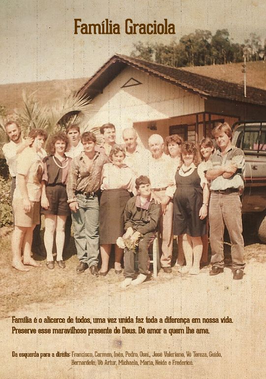 Grupo de pessoas posando em frente a uma casa de madeira, com a inscrição “Família Graciola” acima dela.