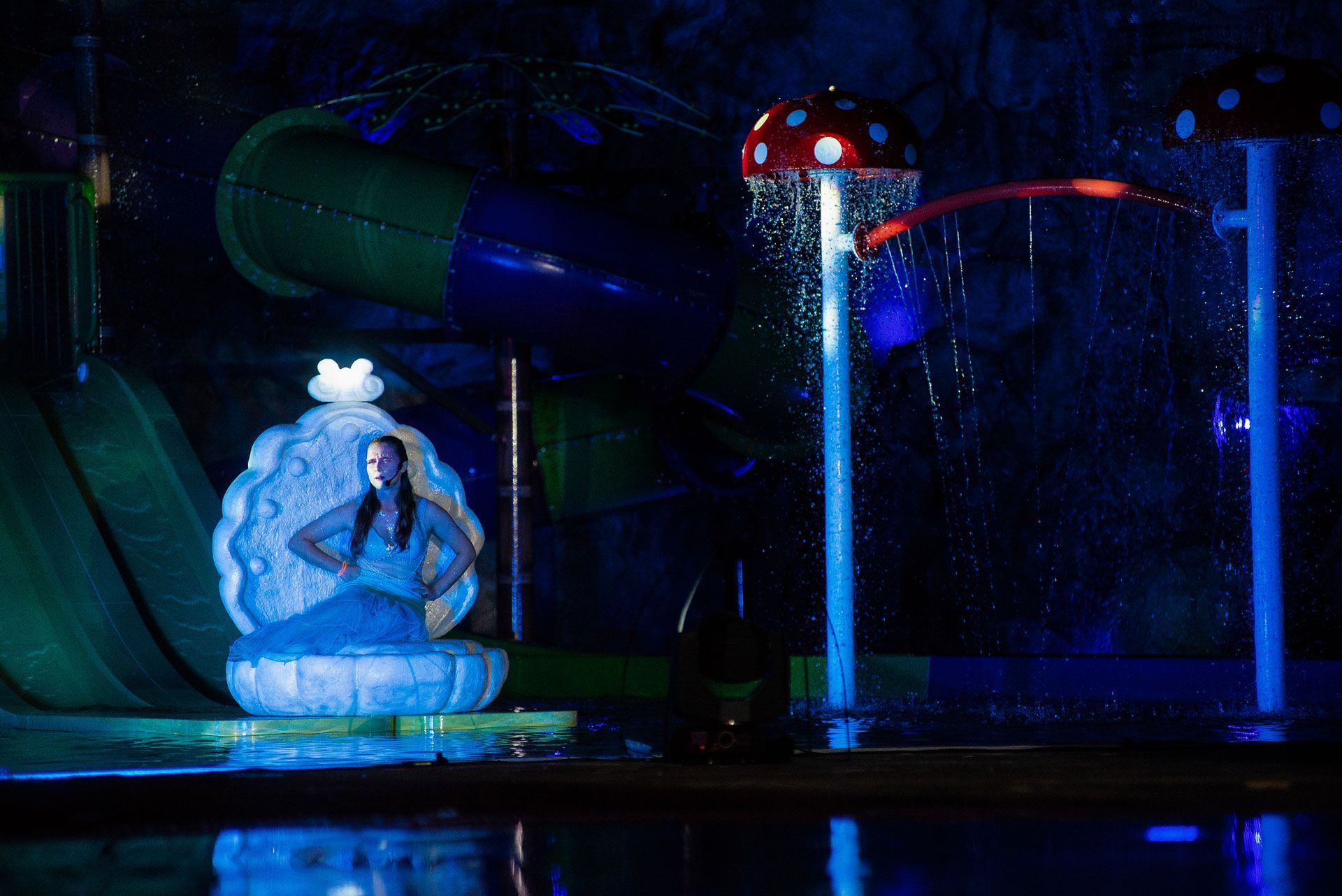 Espetáculo aquático noturno com uma estátua de sereia iluminada em azul e fontes coloridas jorrando água em um palco escuro.