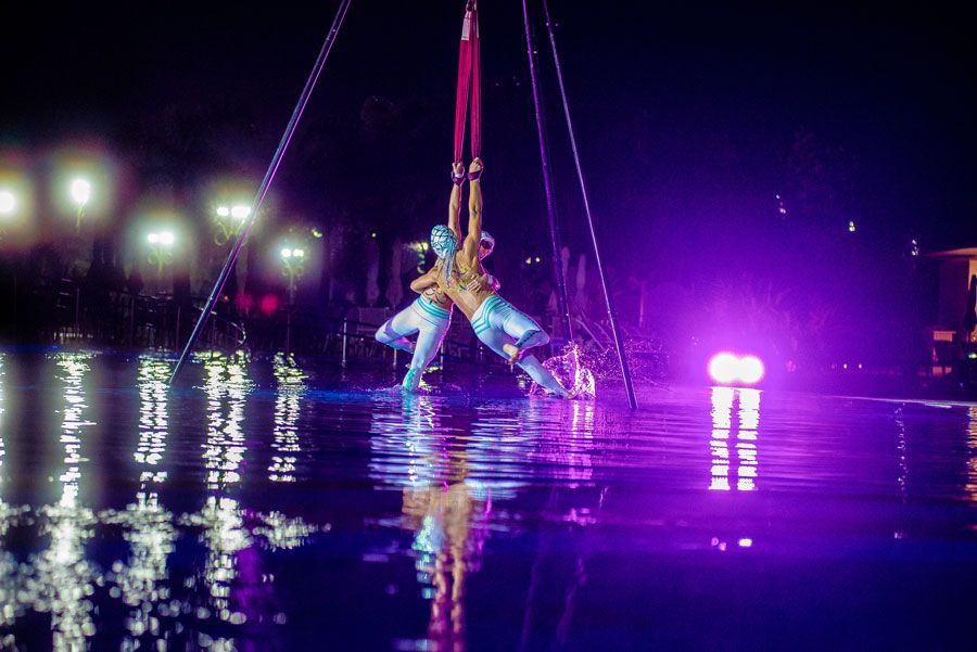 Artista aérea suspensa sobre a água à noite, com luzes roxas do palco refletidas na superfície.