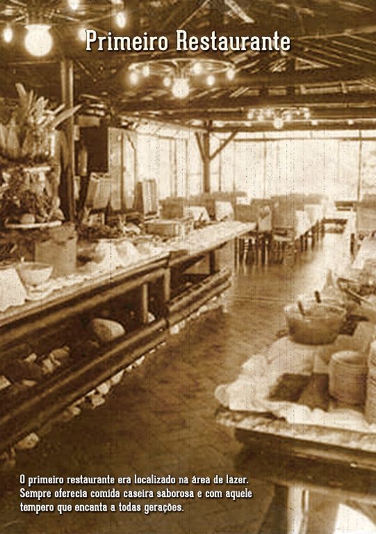 Interior em tons sépia do primeiro restaurante, com mesa comprida, vigas de madeira e luminárias pendentes.