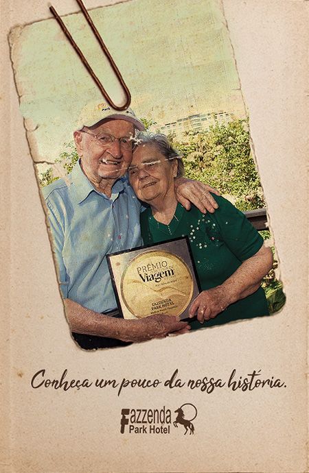 Elderly couple smiling and hugging, holding a plaque, with “Cozinha um pouco da sua história” text below.