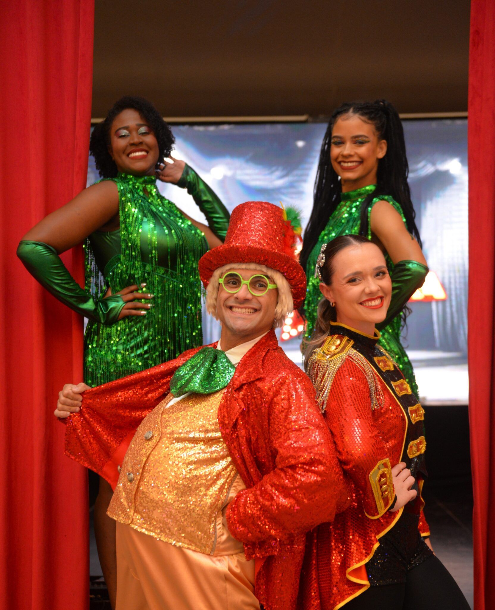 Quatro artistas com trajes brilhantes em vermelho, verde e dourado posando no palco, sorrindo com as mãos na cintura.