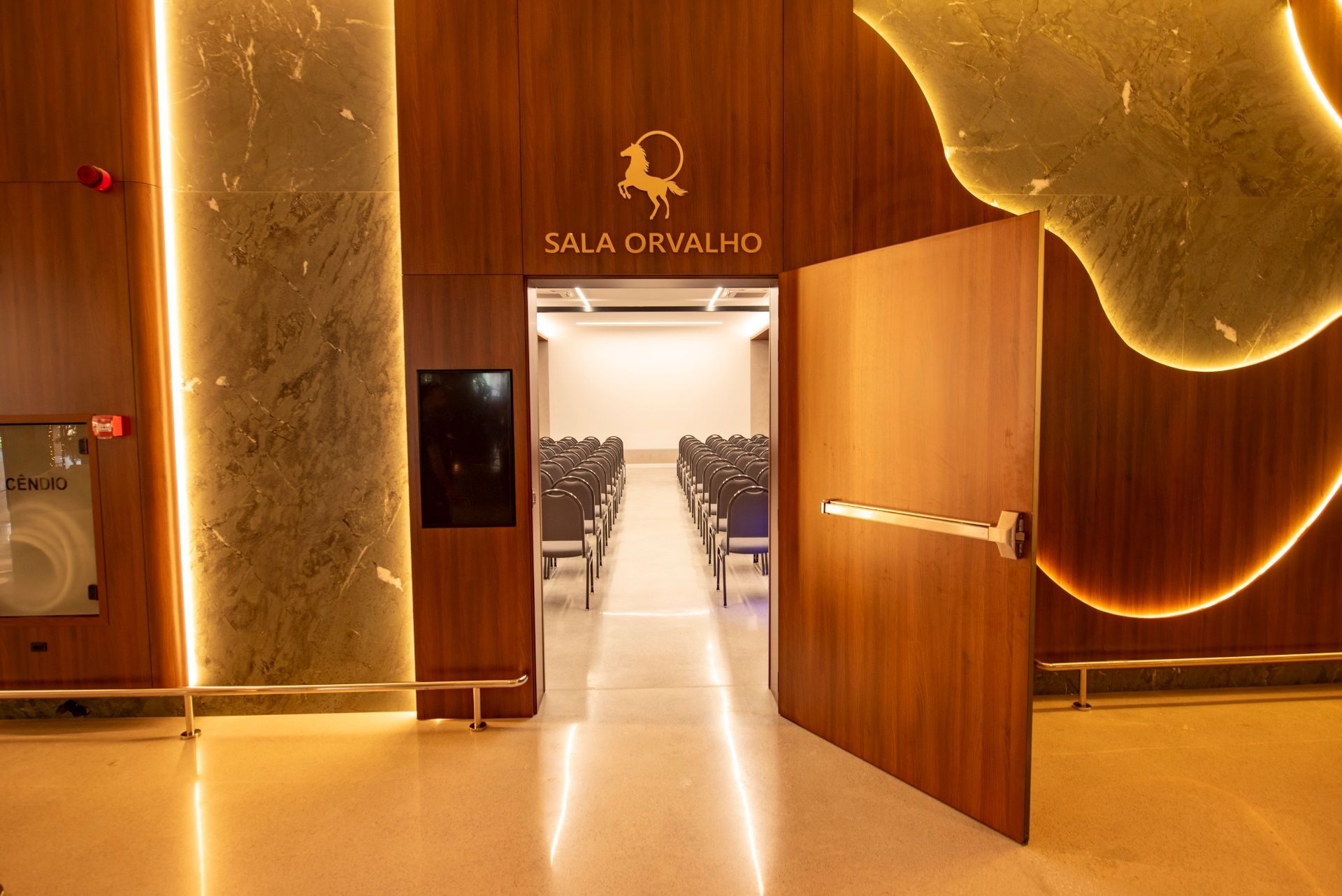 Entrada com painéis de madeira para uma sala de reuniões "Sala Oratório" com fileiras de cadeiras no interior.