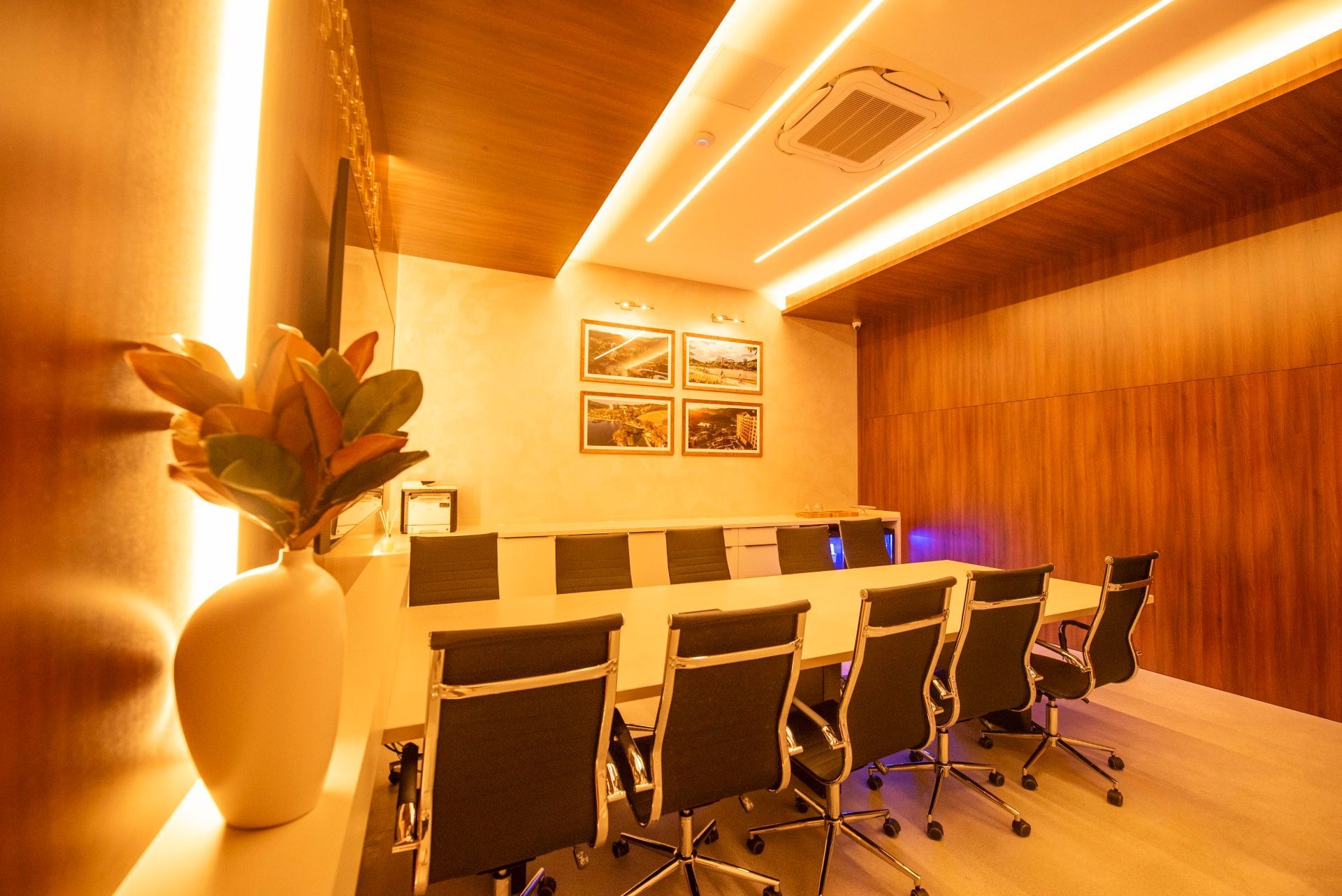 Sala de conferências moderna com mesa comprida, cadeiras pretas, obras de arte na parede e iluminação aconchegante com painéis de madeira.