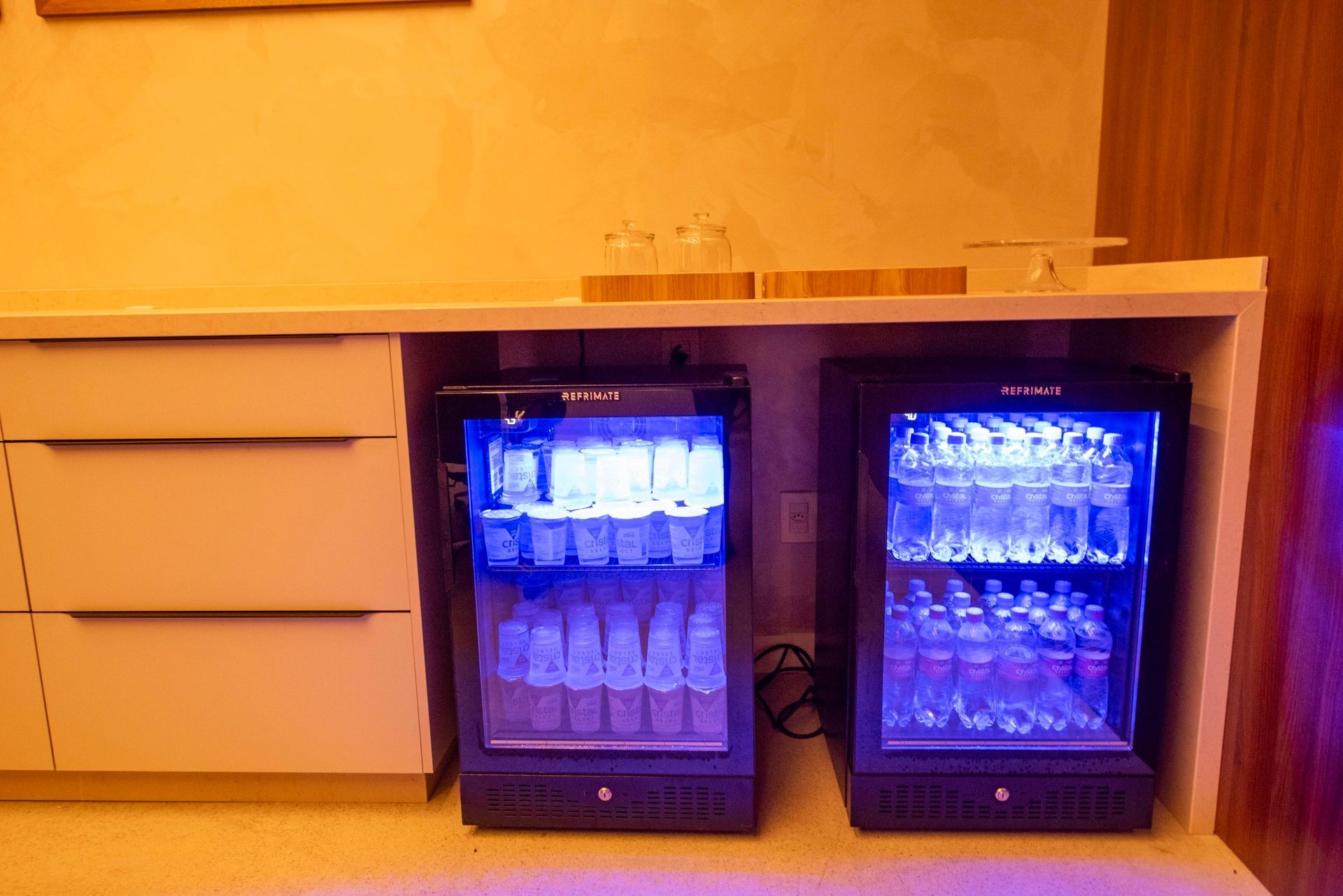 Duas geladeiras pretas para bebidas, iluminadas em azul, sob um balcão, em uma sala com gavetas bege.