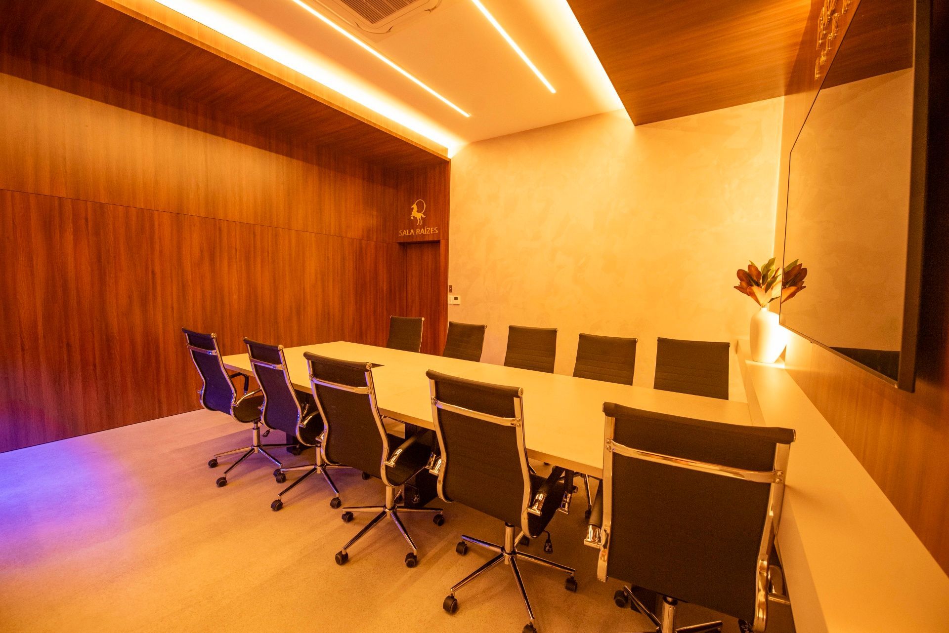 Sala de conferências moderna com uma mesa comprida, cadeiras pretas, paredes revestidas de madeira e iluminação aconchegante no teto.