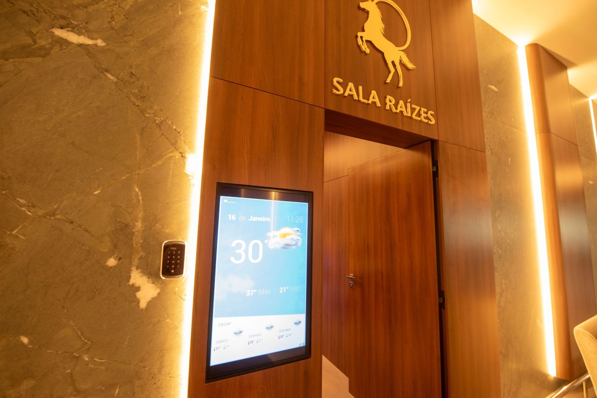 Entrada com painéis de madeira e a inscrição “SALA RAÍZES”, com uma tela digital iluminada ao lado da porta.