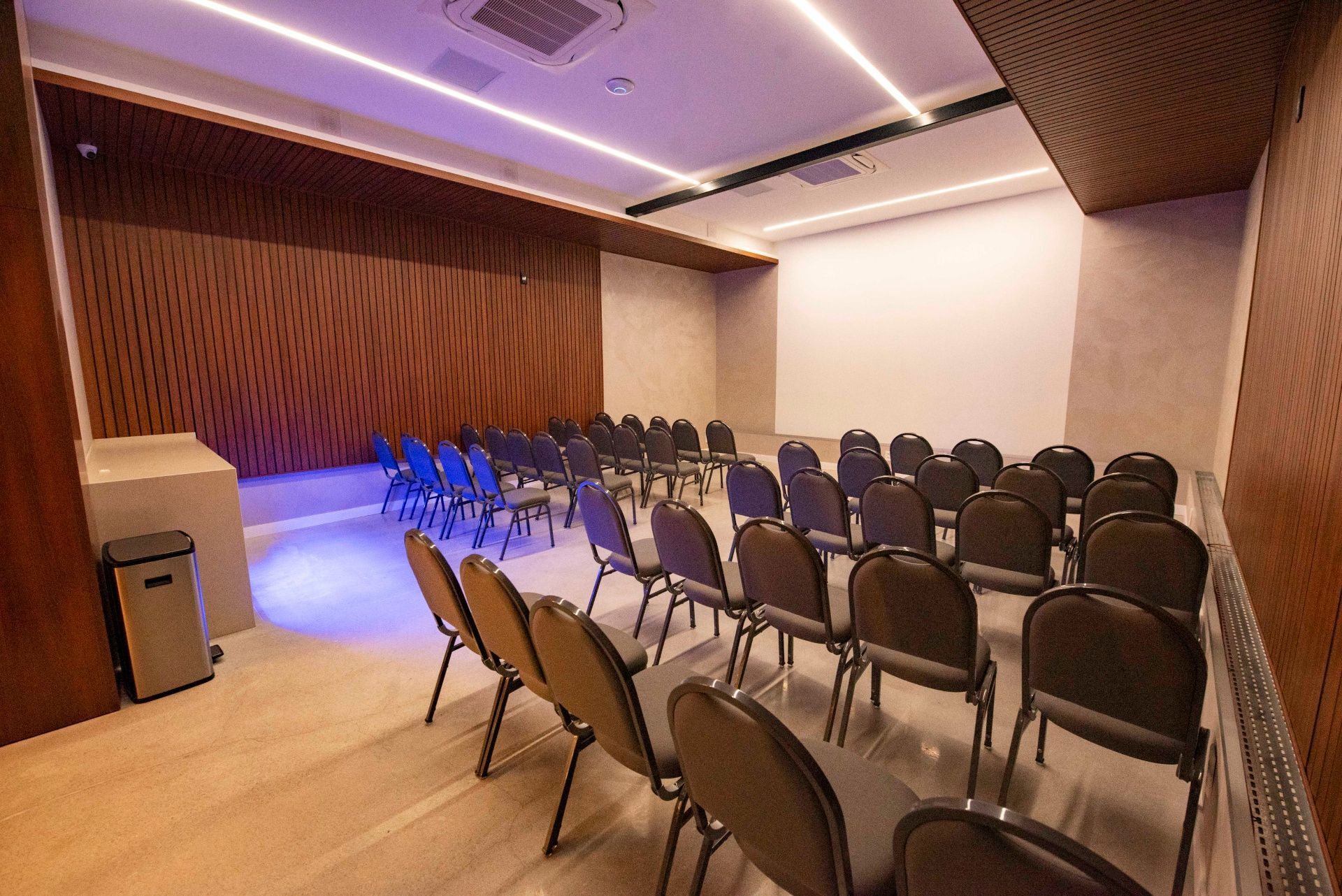 Sala de conferências vazia com fileiras de cadeiras, paredes revestidas de madeira e iluminação suave no teto em tom roxo.