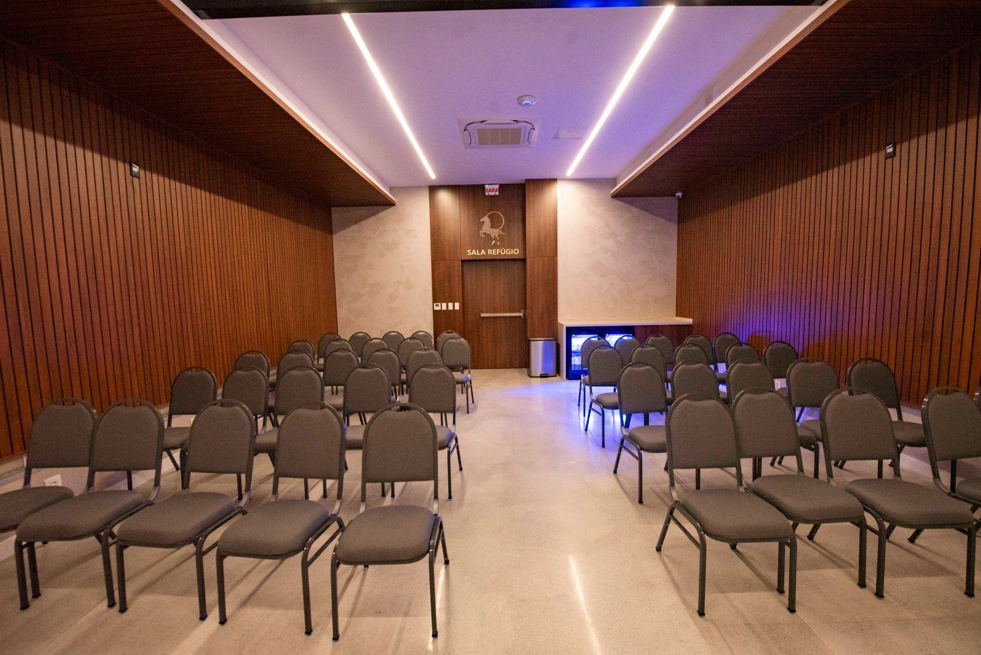 Sala de conferências moderna e vazia, com fileiras de cadeiras cinzentas, paredes revestidas de madeira e iluminação suave no teto.