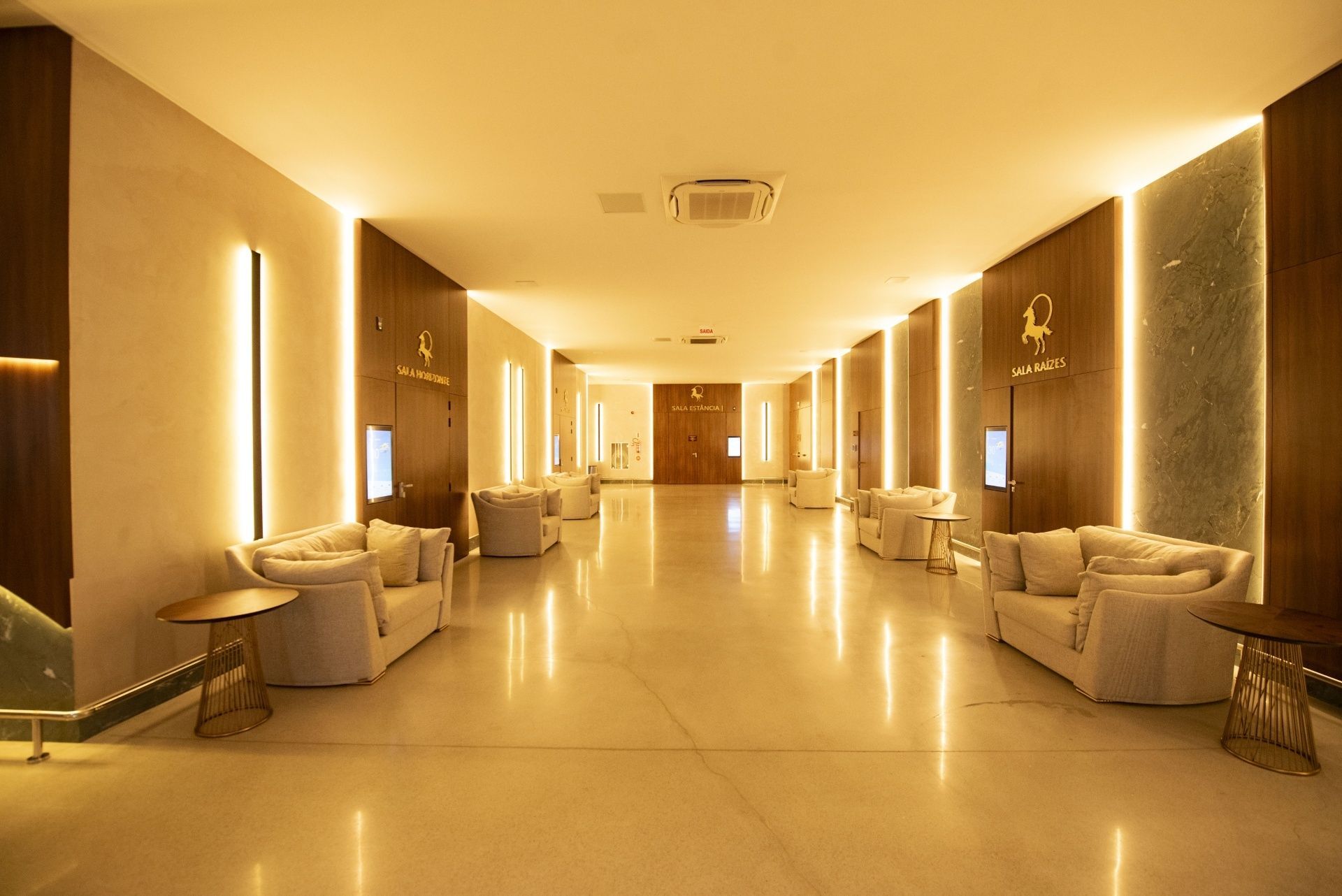 Corredor de hotel com iluminação aconchegante, poltronas e apliques de parede em ambos os lados.