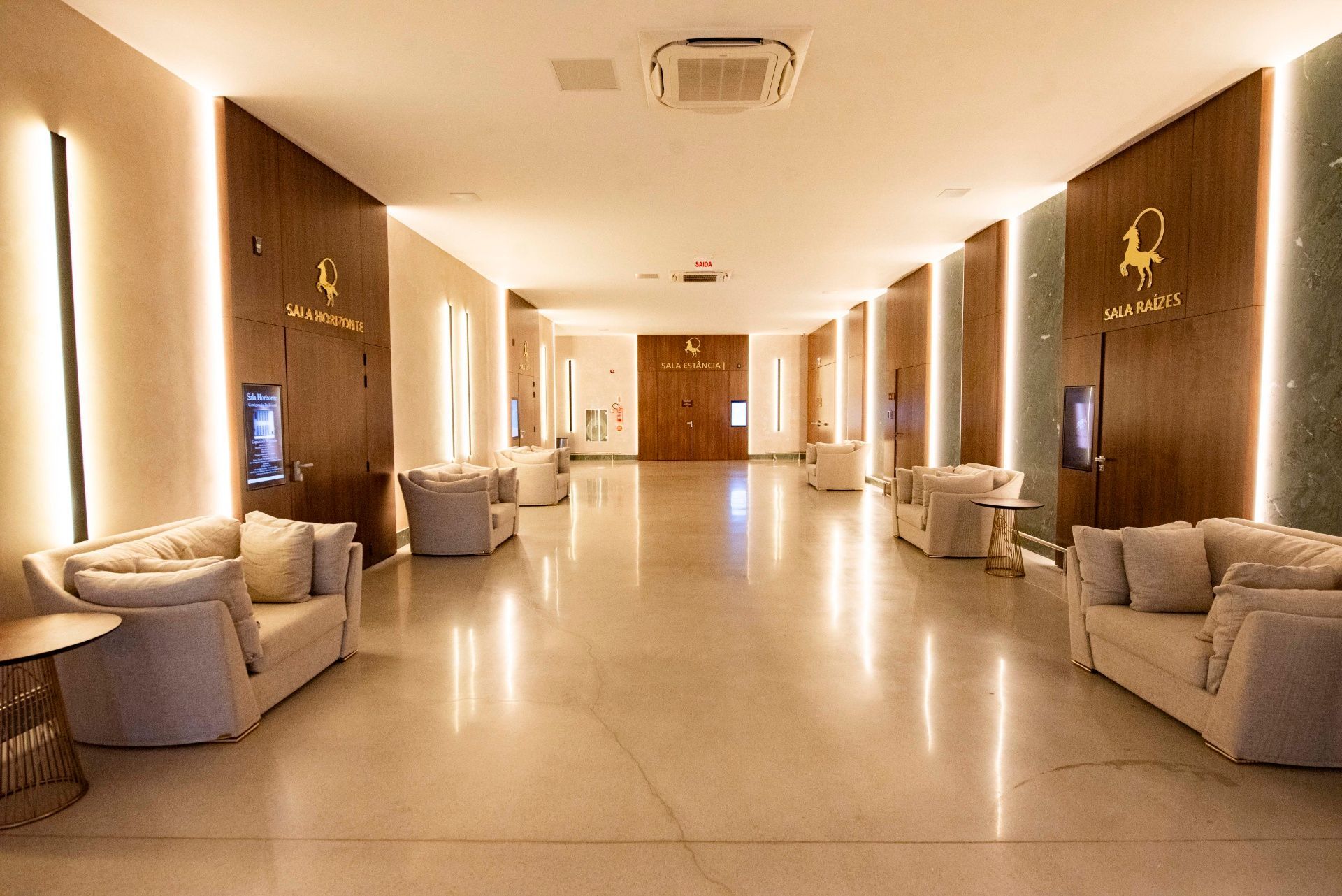 Corredor espaçoso do hotel com paredes cor creme, assentos bege e iluminação aconchegante.