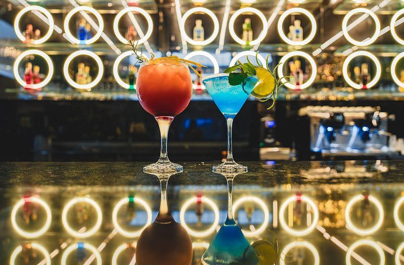 Dois coquetéis coloridos em um balcão de bar, com luzes circulares brilhantes refletidas no fundo e na superfície.