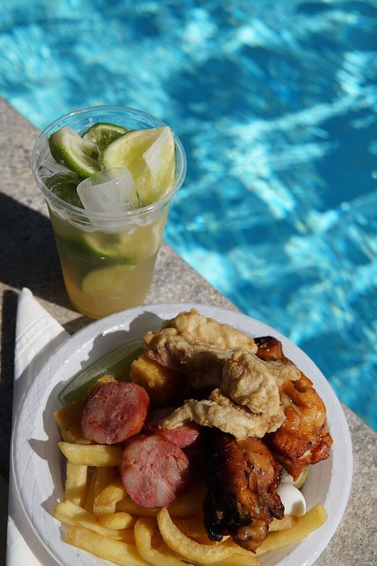 Prato de comida frita e batatas à beira da piscina, acompanhado de uma bebida de limão com gelo e fatias de limão.