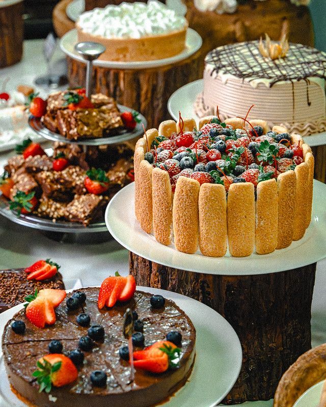 Diversos bolos e sobremesas decorados sobre uma mesa, incluindo bolos com cobertura de morango e recheados com frutas vermelhas.