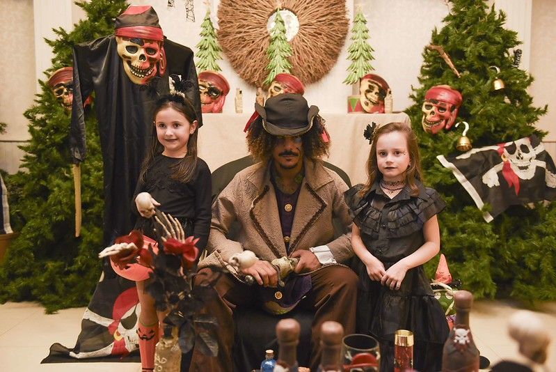 Grupo fantasiado posando em frente a uma decoração de Halloween com árvores, caveiras e enfeites assustadores.