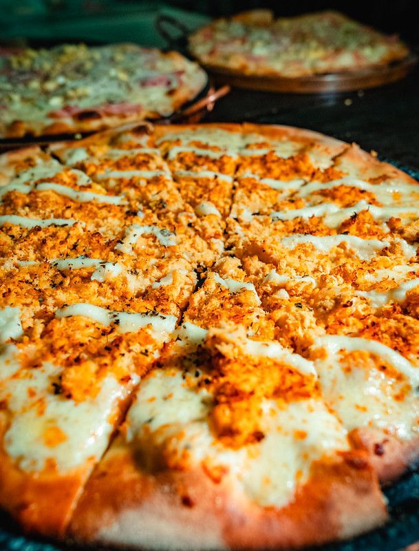 Close-up de uma pizza fatiada e recheada com queijo e tempero de laranja sobre uma mesa escura, com outras pizzas desfocadas ao fundo.
