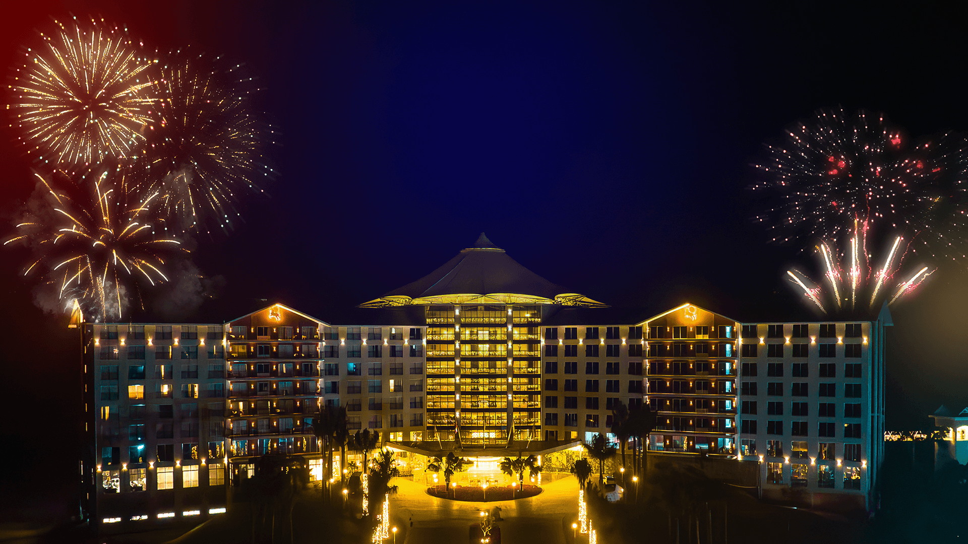 Hotel iluminado em tons dourados à noite, com fogos de artifício explodindo em ambos os lados.