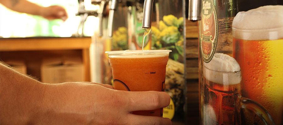 Mãos segurando um copo de suco de laranja ao lado de um smoothie de frutas e uma bebida gelada alta em uma mesa.