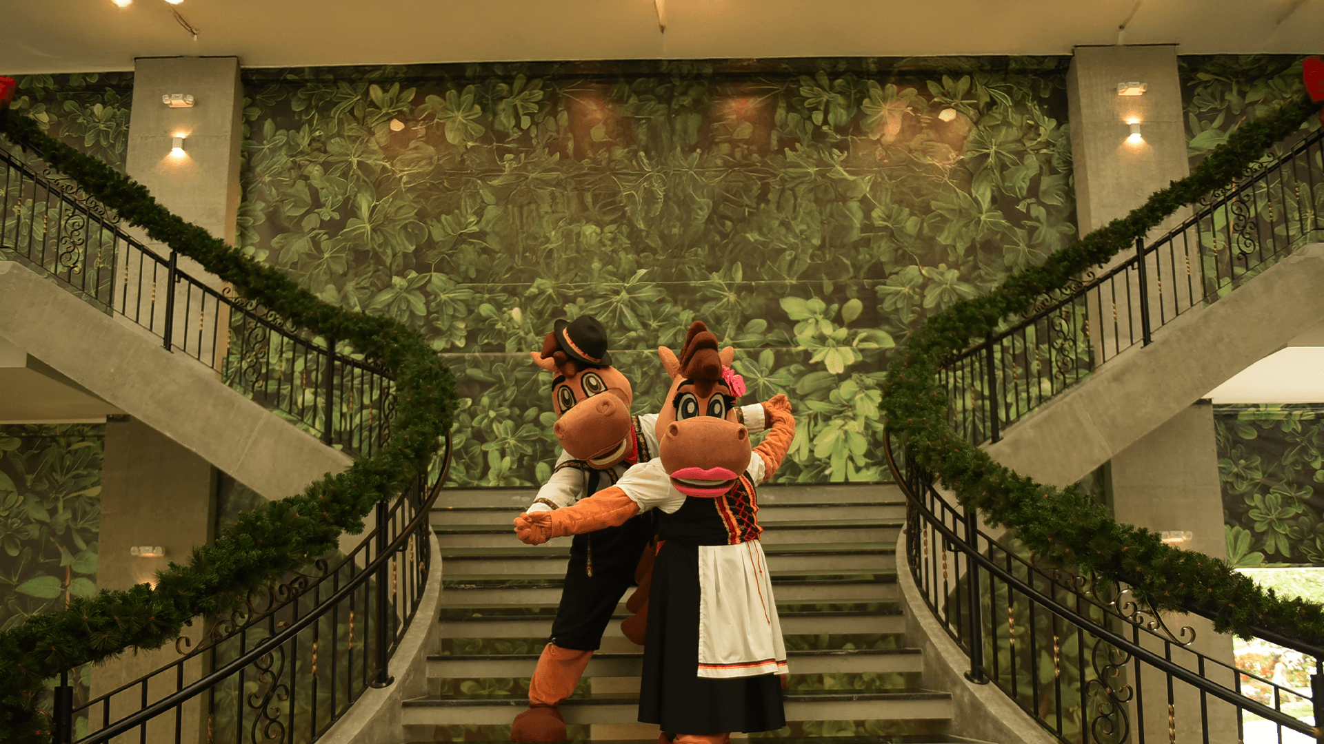 Duas figuras vestidas a caráter estão em uma escadaria em um grande saguão com um mural floral verde ao fundo.