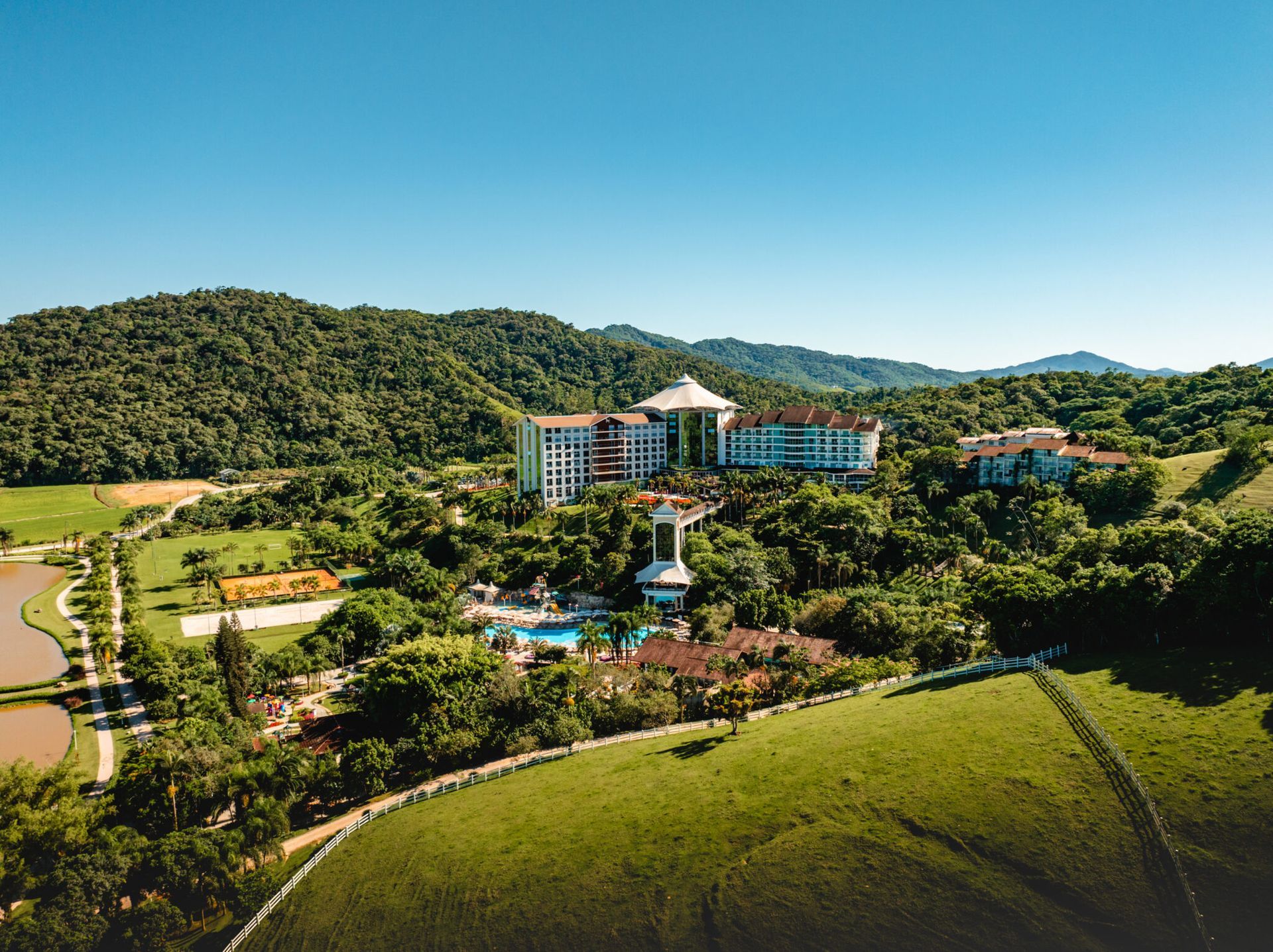 Vista aérea de um resort tropical com edifícios de telhados azuis, piscina e colinas verdejantes sob um céu limpo.