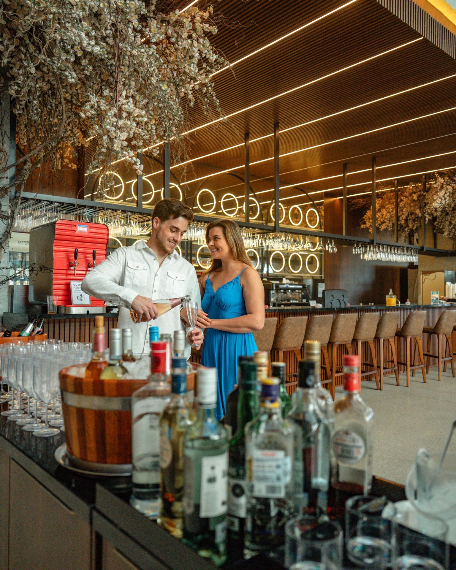 Casal preparando drinques em um bar animado, com garrafas em primeiro plano e luzes pendentes aconchegantes acima.