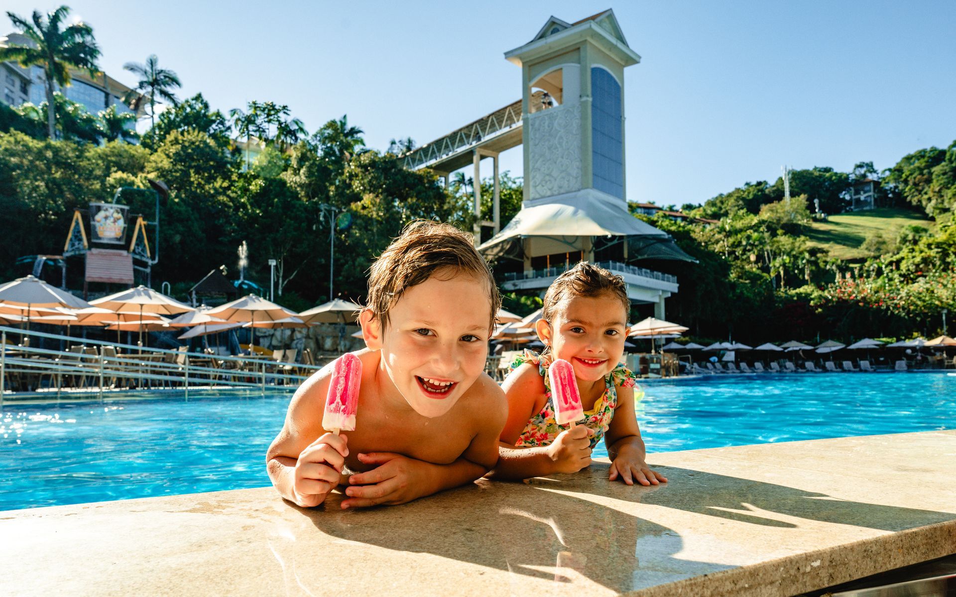Duas crianças sorrindo à beira da piscina em um resort ensolarado com palmeiras e uma torre ao fundo.