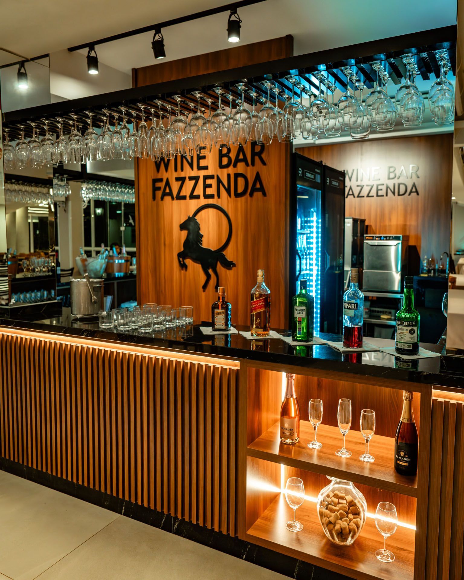 Balcão de bar de vinhos com taças penduradas, placa laranja e prateleiras iluminadas em um interior moderno.