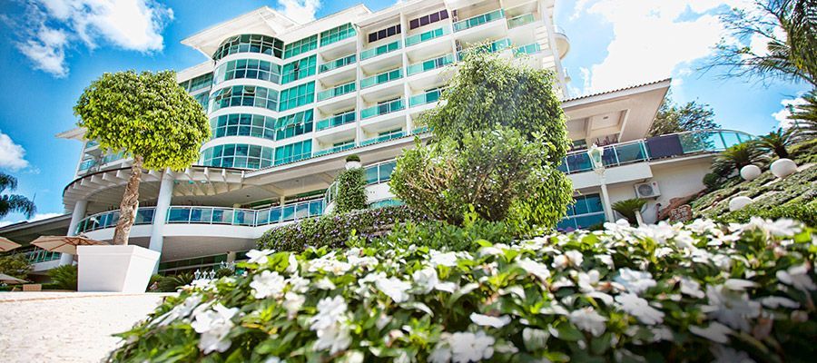 Prédio de apartamentos moderno e branco com varandas e paisagismo exuberante sob um céu azul.