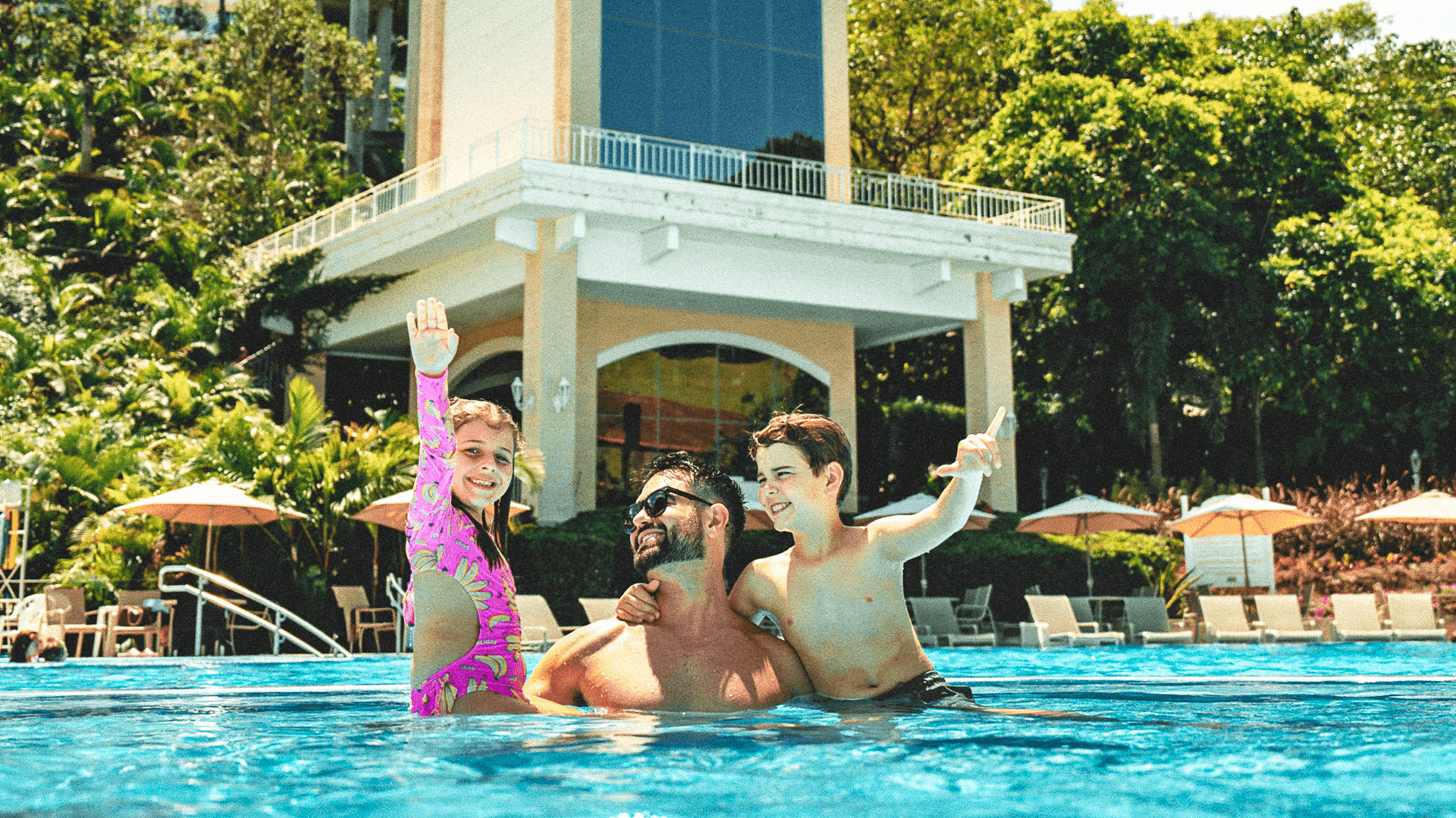 Pessoas se refrescando na piscina e posando para fotos em frente a um prédio de um resort.