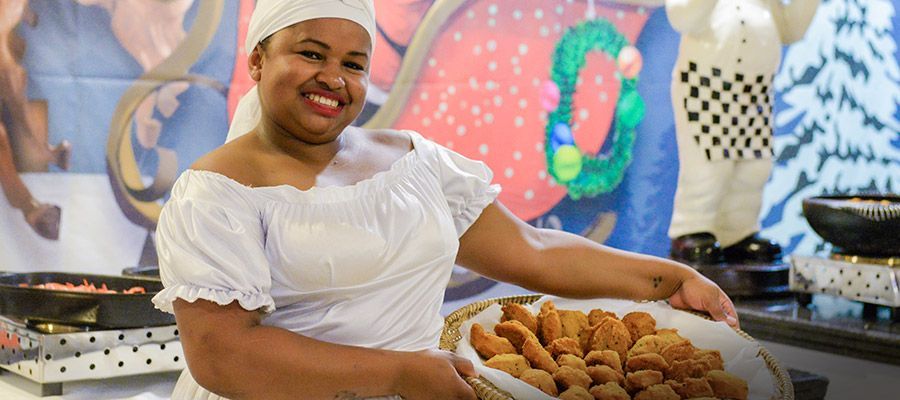 Chef de uniforme branco sorrindo enquanto apresenta uma bandeja de doces em uma cozinha colorida.