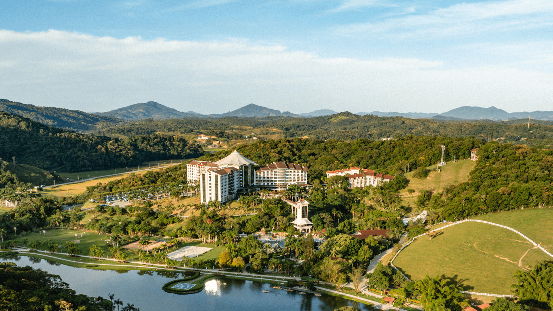 Hotel resort moderno com fachadas brancas e vermelhas, átrio central de vidro e jardim paisagístico sob céu azul.