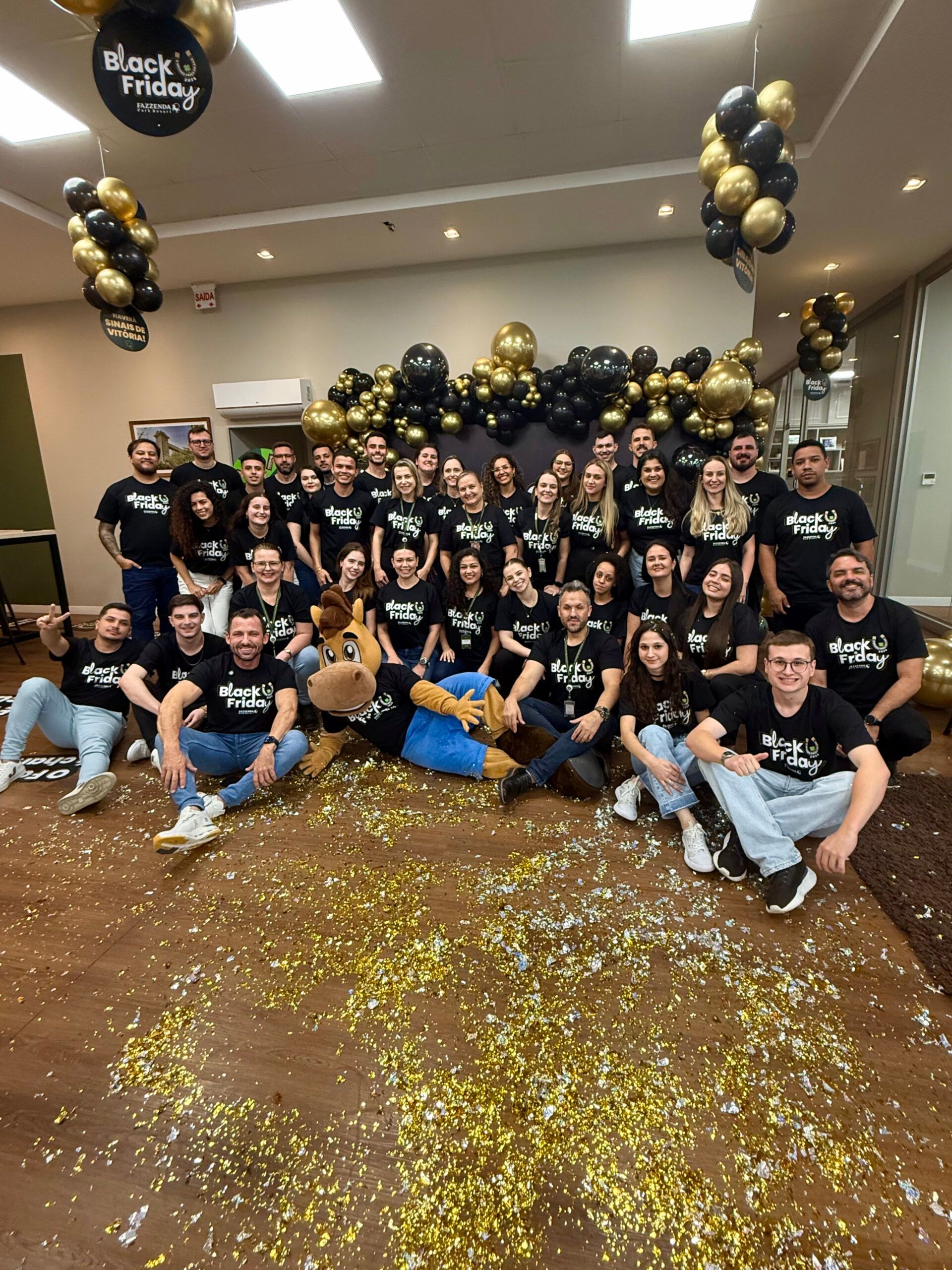 Um grande grupo posando em um ambiente interno, em meio a confetes dourados e balões pretos e dourados, para celebrar um evento da equipe.