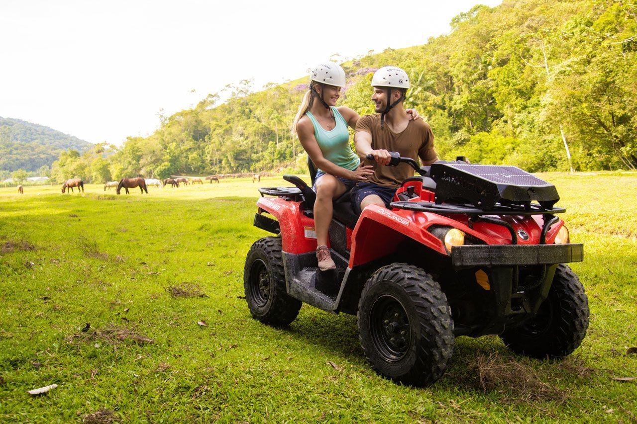 Duas pessoas conduzindo um quadriciclo vermelho por um campo gramado com colinas ao fundo.