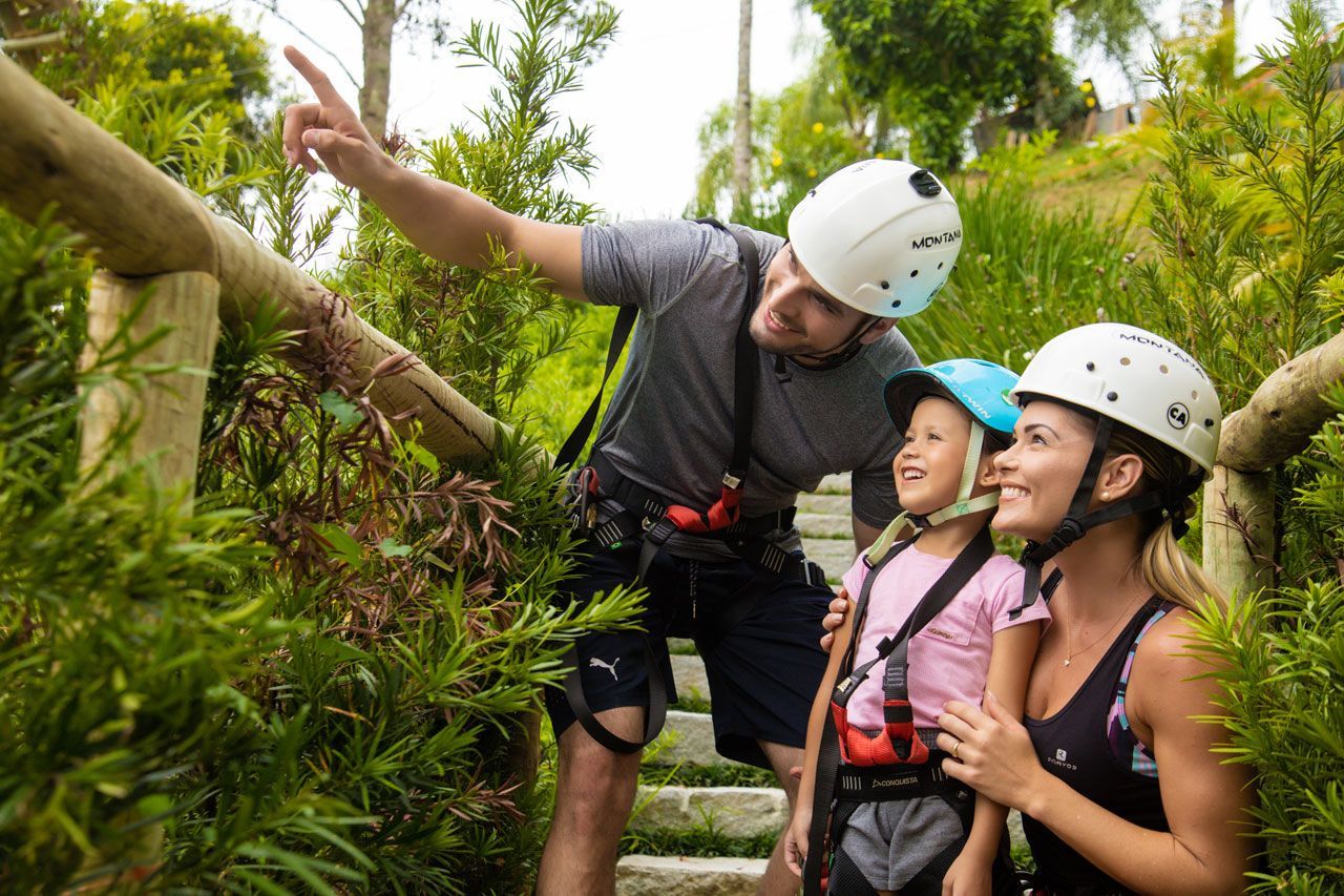 Três alpinistas de capacete e arnês, sorrindo, em uma plataforma de tirolesa na selva.