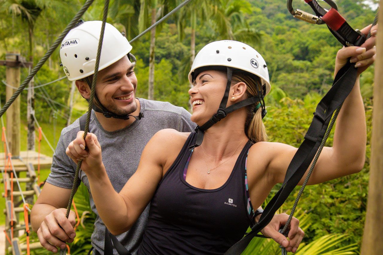 Duas pessoas de capacete sorrindo em uma tirolesa sobre uma floresta verdejante.