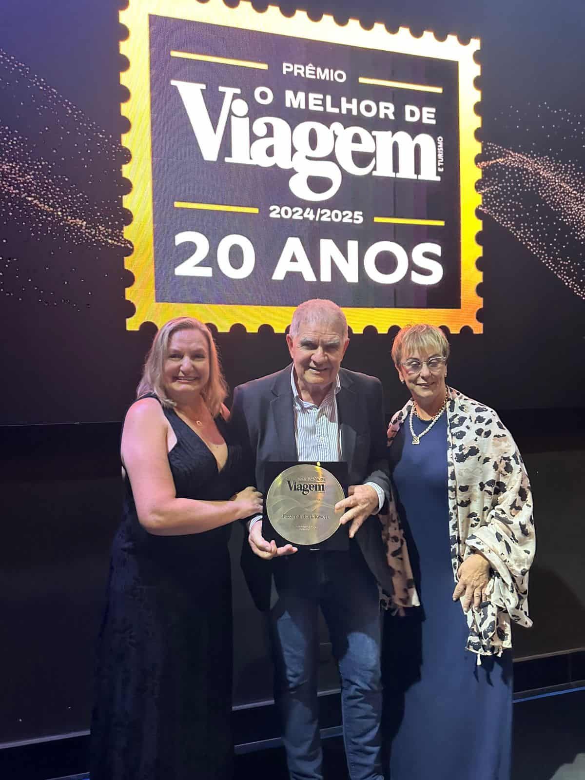 Três pessoas posam com troféu diante do painel “Melhor de Viagem 2024/2025 – 20 anos”.