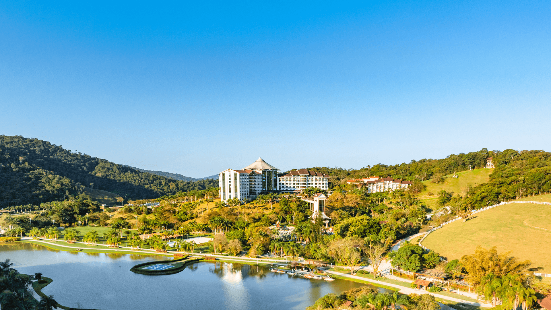 Resort à beira do lago em uma encosta ensolarada com água azul, colinas verdejantes e edifícios sob um céu claro.