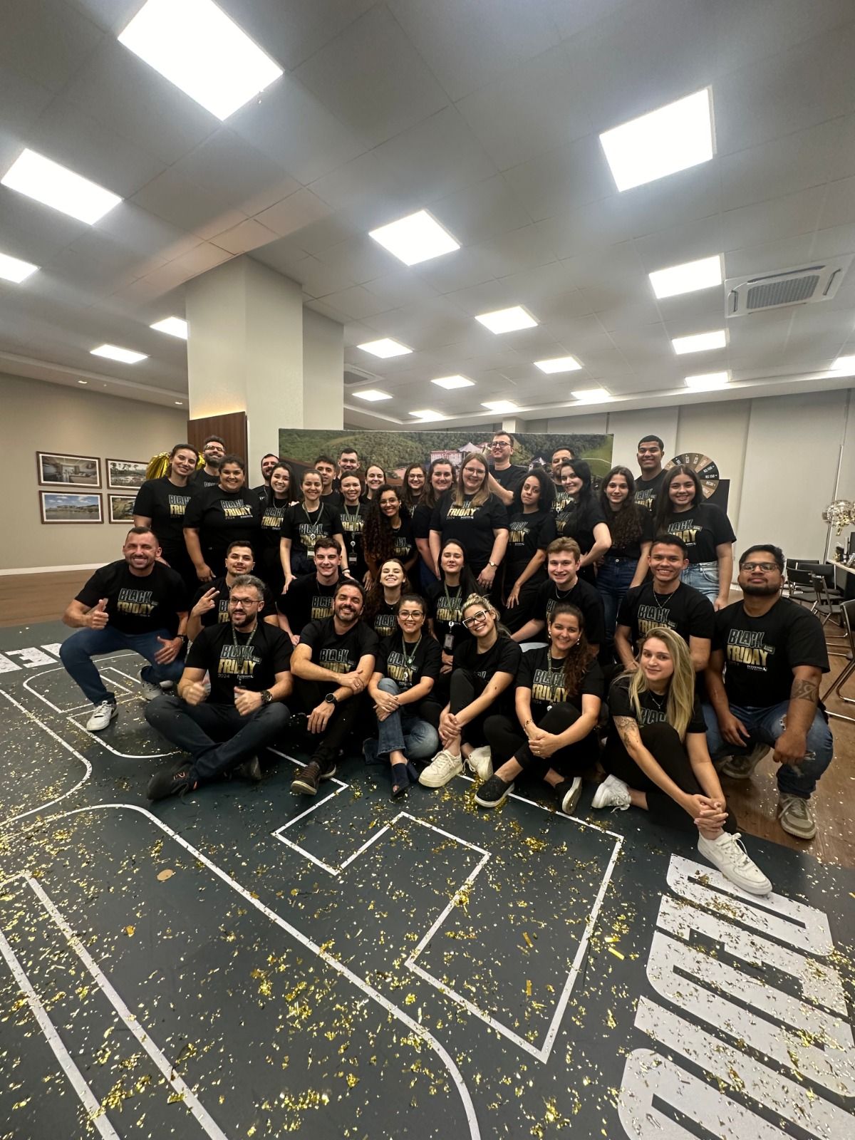 Um grande grupo posando em uma sala de conferências com carpete estampado e luzes brilhantes no teto.