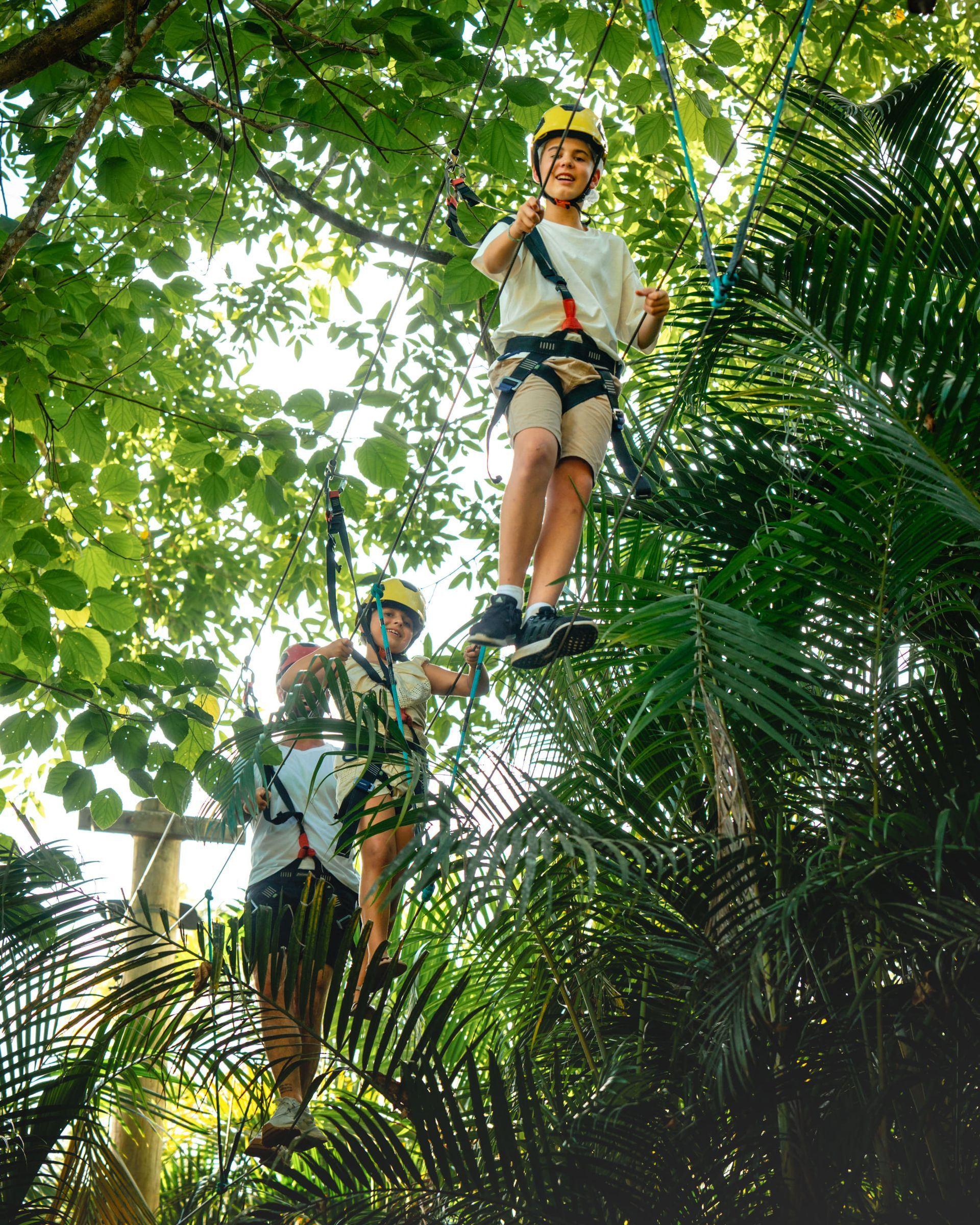 Três alpinistas com capacete, sorrindo, em um percurso de cordas em uma floresta exuberante.