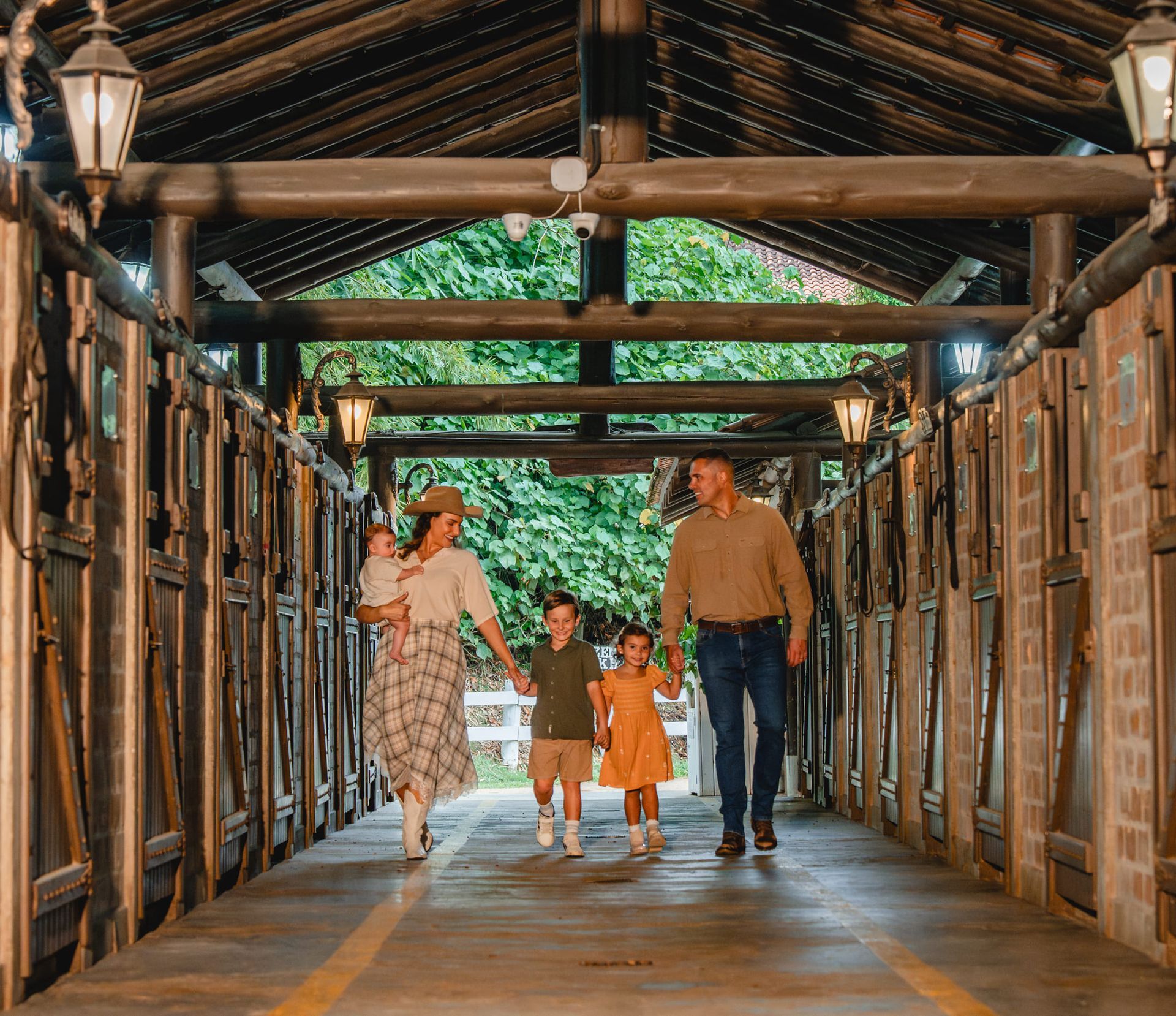 Família caminhando por uma passarela de madeira coberta, com iluminação aconchegante e árvores verdes ao fundo.