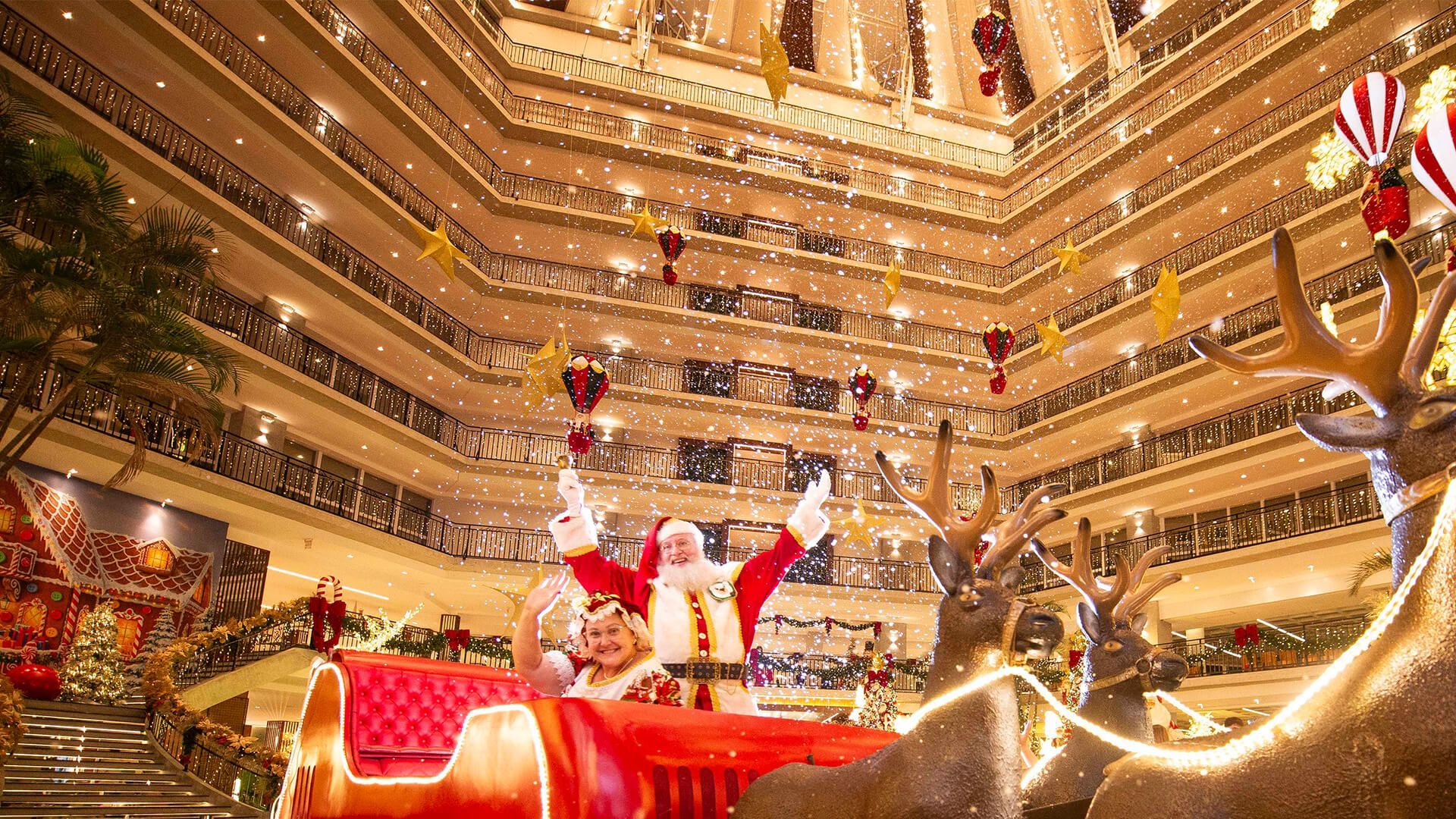 Papai Noel no saguão de um grande hotel com uma árvore de Natal decorada e confetes caindo.
