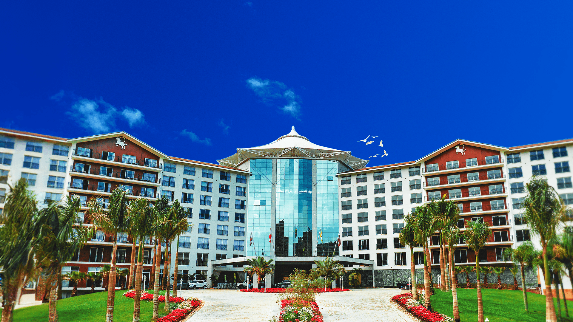 Hotel resort moderno com fachadas brancas e vermelhas, átrio central de vidro e jardim paisagístico sob céu azul.