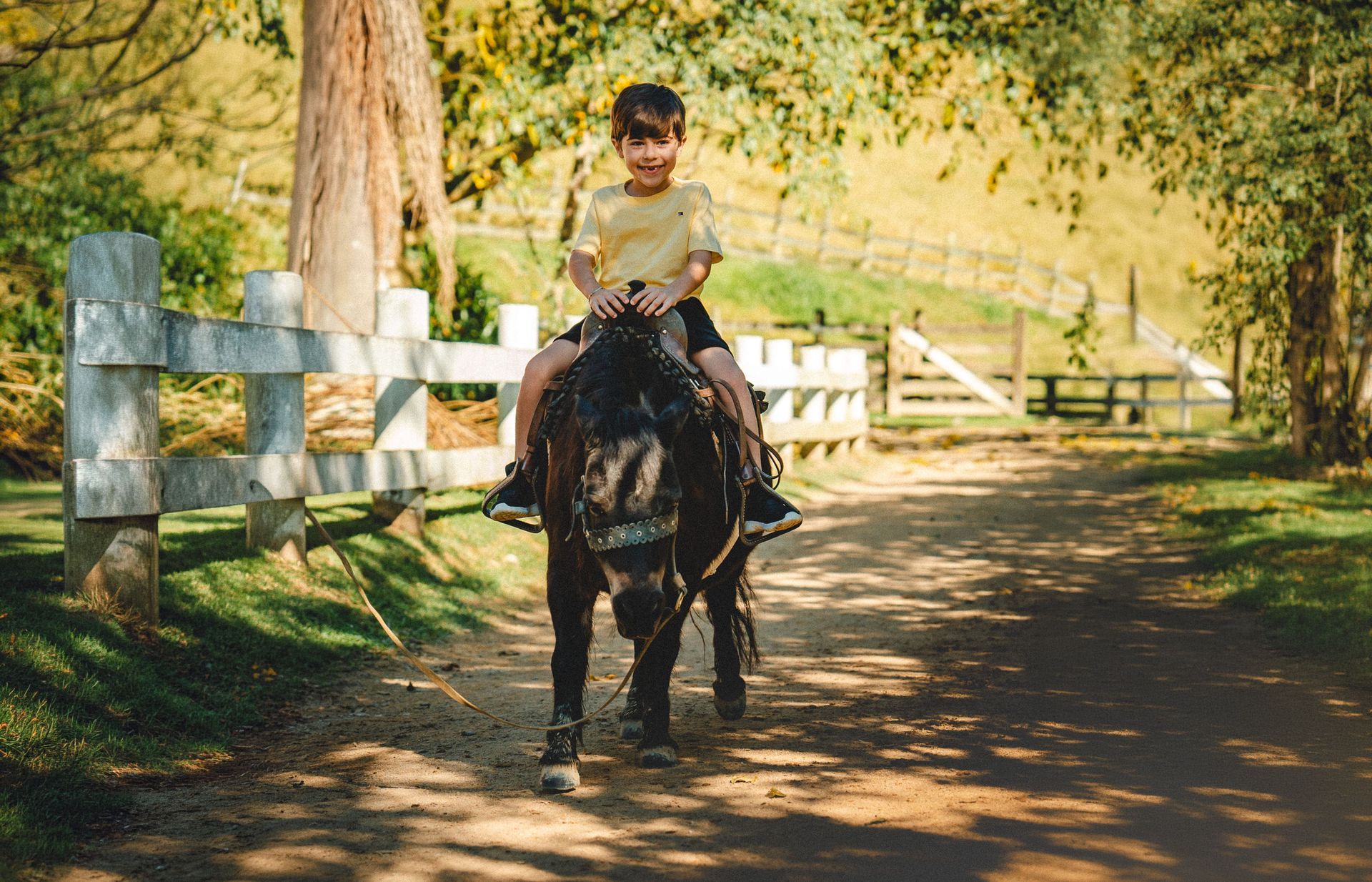 Criança a cavalo, sorrindo e com um braço levantado em uma área ensolarada para equitação ao ar livre.